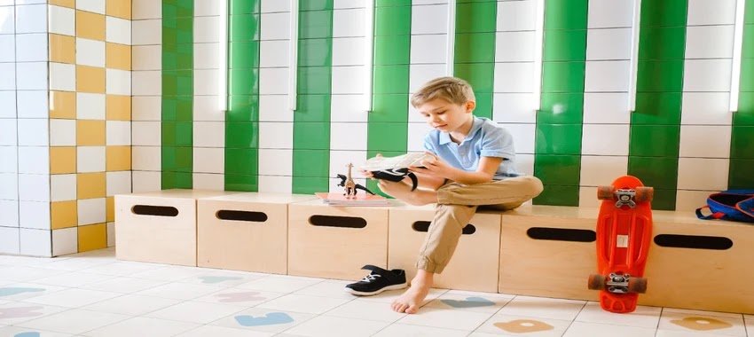 A young boy sitting on a wooden bench in a tiled locker room, adjusting his shoe while barefoot on one foot, with a skateboard nearby.